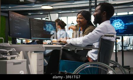 Joyous technician with disability and coworker chatting while coding AI algorithms debugging software issues. Upbeat tech startup office IT expert in wheelchair and colleague using computer, camera A Stock Photo