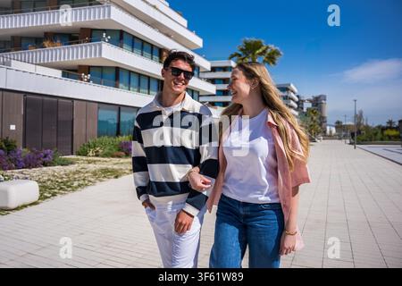 Young couple walking arm in arm and smiling past modern apartment buildings on a sunny day Stock Photo