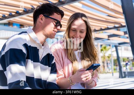 Young caucasian couple sitting closely together, smiling as they look at a smartphone screen on a sunny day Stock Photo