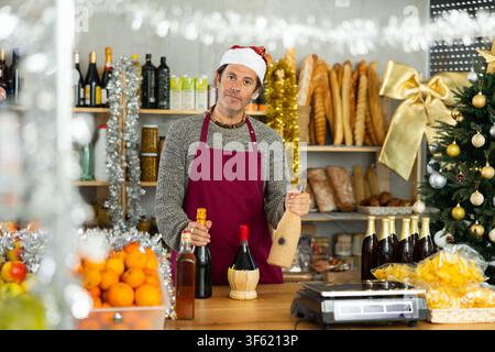 Friendly adult male shop seller posing behind counter with alcoholic drinks, Christmas goods in market Stock Photo