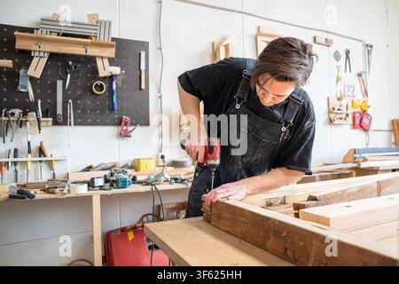 Woodworker technician using handheld cordless electric driver to fasten long screws in wooden frame. Stock Photo