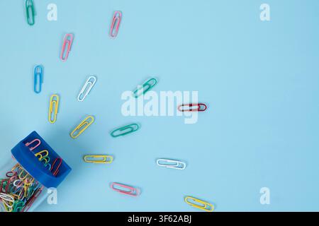 Colorful paper clips scattered on a light blue background with a small transparent jar filled with paper clips Stock Photo