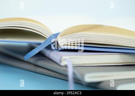 Open notebook with blank pages and ribbon bookmark resting on a stack of closed notebooks on a light blue background Stock Photo