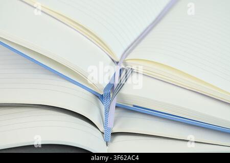 Open notebook with blank pages and ribbon bookmark resting on a stack of closed notebooks on a light blue background Stock Photo