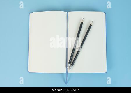 Two black pencils arranged on blank pages of an open notebook on a light blue background, top view Stock Photo