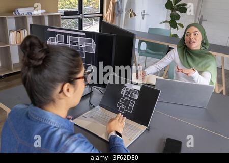 Diverse female coworkers collaborating on design in office, reviewing laptop floorplan and monitors. Architecture, planning, blueprint, rendering, collaboration, teamwork, modern Stock Photo