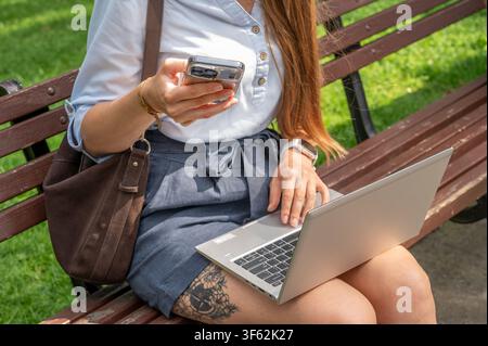 Businesswoman sitting on park bench holding smartphone and using laptop outdoors on sunny day. Young female office worker multitasking during lunch br Stock Photo