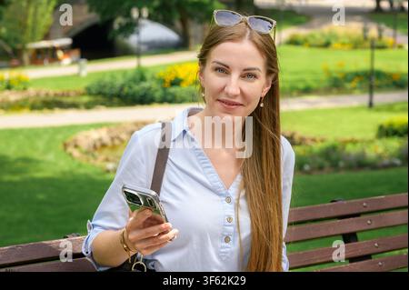 Portrait of smiling female student sitting on bench in summer park with smartphone in hand. Young woman using mobile phone outdoors Stock Photo
