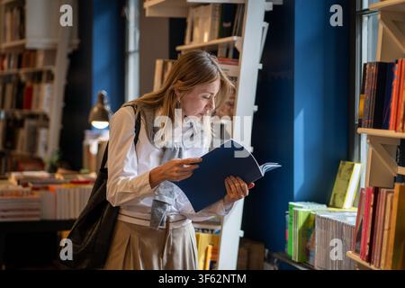 Intelligent woman booklover reading book in cozy bookshop, flipping through pages of publication. Stock Photo