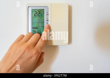 Hand adjusting a digital thermostat set to 22 degrees on a modern wall mounted climate controller. Smart home technology, indoor comfort and energy Stock Photo