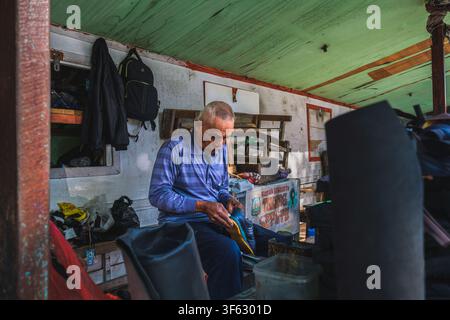 Balikpapan, Indonesia - June 18 2026. A senior cobbler sitting inside his street-side workshop stall, completely focused on  repairing a boot. Stock Photo