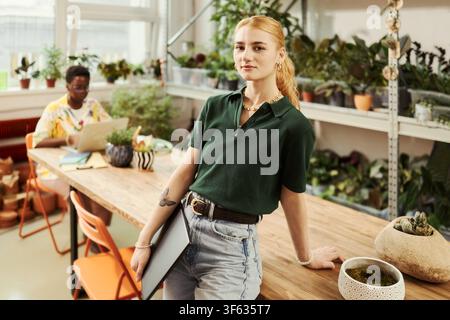 Portrait of young Caucasian woman holding laptop in creative plant filled office while African American colleague works behind. Ideal for modern workplace diversity marketing Stock Photo