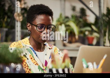 African American young woman wearing glasses working on laptop in creative office, focusing on screen. Image suitable for remote work and diverse business marketing Stock Photo