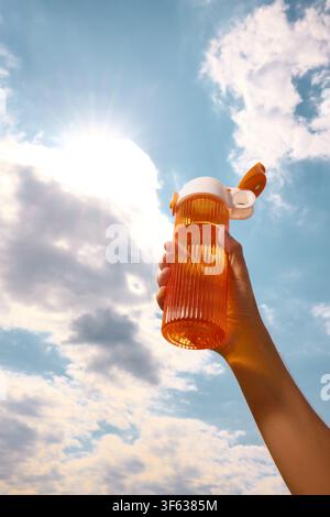 Orange sports bottle held high against sunlit clouds during outdoor workout Stock Photo