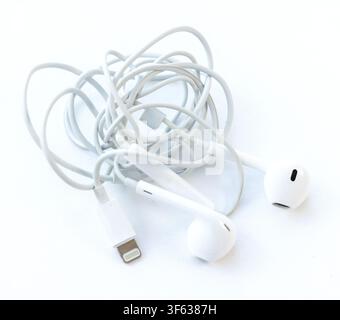 A tangled pair of white earphones with a charging connector lies in a messy heap on a white surface Stock Photo