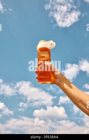 Hand holding orange sports bottle high against sunny cloudscape Stock Photo