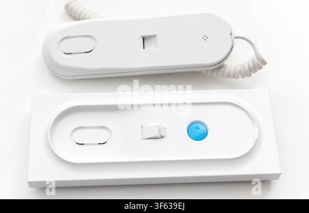 A white intercom handset with a coiled cord is placed next to its matching wallmounted base unit featuring a blue key button Stock Photo