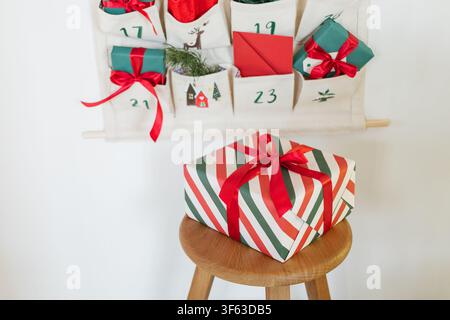 Close-up of part of a Traditional Christmas advent calendar with assorted wrapped gifts hanging on a white wall next to a wooden stool with a larger wrapped Christmas gift Stock Photo