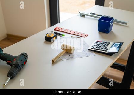 Construction tools including a drill, tape measure, and wooden mallet arranged on the table alongside architectural plans.  Stock Photo