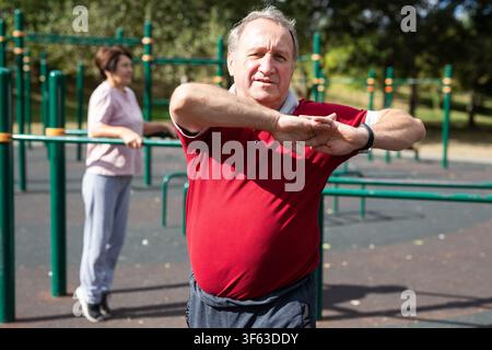 Staying after retirement. Happy joyful mature retired sportsman wearing sportswear doing side stretching exercises with arm over his head, exercising Stock Photo