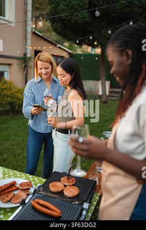 Diverse women friends enjoying backyard barbecue and using smartphone Stock Photo