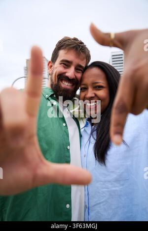 Happy multiracial couple taking a photo framing with hands in a city park Stock Photo