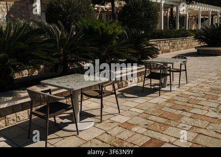 Modern outdoor cafe terrace with empty tables and chairs surrounded by lush greenery. Peaceful hospitality setting in warm summer sunlight. Stock Photo