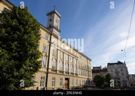 serene historical clock tower in urban setting with large tree and other buildings visible from low angle, looking up at the structure with stone and Stock Photo