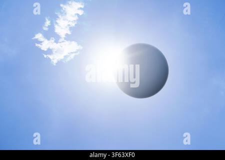 A partial solar eclipse with bright sunlight emerging from behind a dark spherical moon against a clear blue sky with a soft white cloud. Rare natural Stock Photo