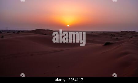 The sun sets in the distance, casting an orange glow over soft dunes in a vast sand desert. The landscape stretches for miles under the evening sky, marking the end of the day. Stock Photo