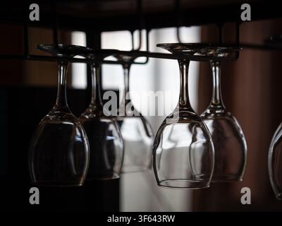 Clear wine glasses are neatly hanging upside down on a metal stemware rack in a bar or restaurant, reflecting ambient light from the interior Stock Photo