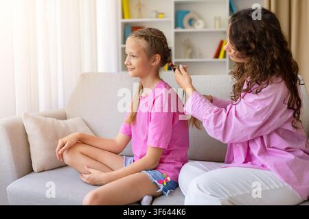 Caring mother braiding her daughters hair while they sit together on sofa Stock Photo
