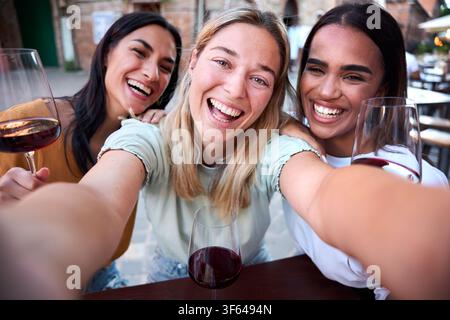 Three happy multi-ethnic friends taking selfie while drinking wine in a restaurant Stock Photo
