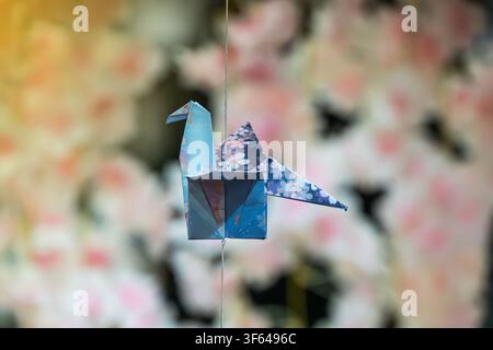Origami crane suspended by wire against floral background, intricate folded paper bird, light blue and dark blue with subtle floral pattern, soft past Stock Photo