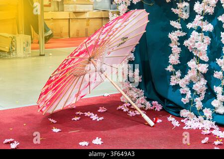 Japanese parasol with vibrant cherry blossom design, pink and white petals scattered, rustic wooden handle, placed on red carpet, teal backdrop, elega Stock Photo