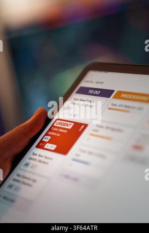 Close Up Hand Holding Digital Tablet Urgent Task Kanban Board Screen Interface. A person works on an agile project management application displaying a Stock Photo