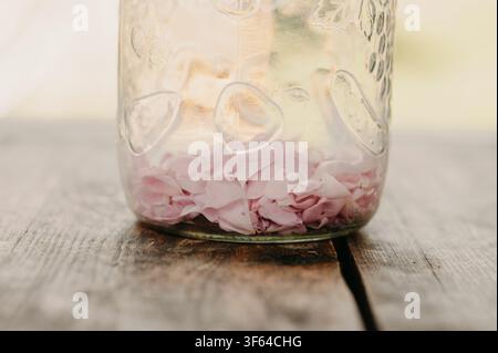 glass jar with pink rose petals on wooden surface, serene outdoor setting, close-up perspective. Stock Photo