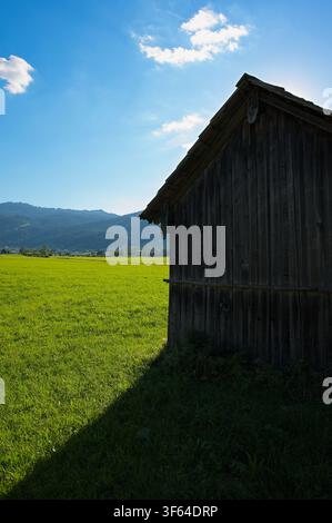 Weathered wooden barn on green sunlit meadow with scenic alpine mountains and blue sky in background Stock Photo