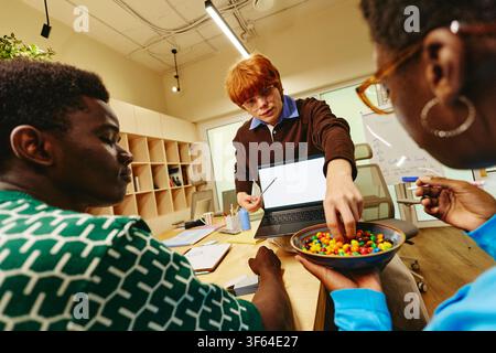 Diverse Colleagues Working on Laptop and Sharing Sweets in Office Stock Photo
