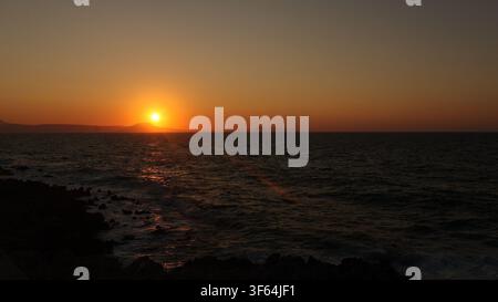 Intense glowing sun casting a strong glare over dark ocean waters during sunset at Rethymno beach, Crete, Greece. Stock Photo