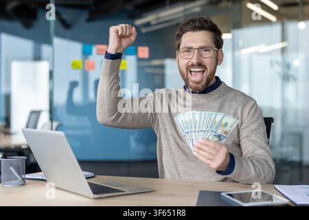 Happy businessman holding cash dollars and raising his fist in celebration while working on a laptop at an office desk, achieving financial success and profit Stock Photo
