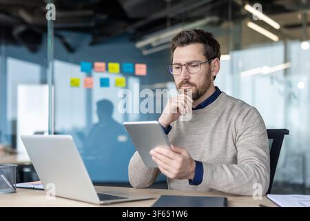 Business man focused on analyzing information on a digital tablet while working with a laptop at a modern office desk, demonstrating concentration and dedication Stock Photo