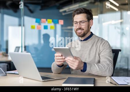 Smiling professional businessman holding a digital tablet while working at his desk with a laptop, reflecting modern office productivity and digital technology Stock Photo