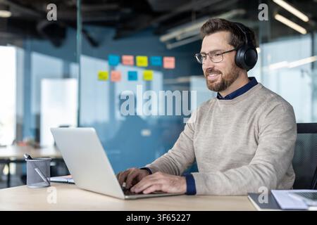 Businessman working remotely from modern office space, smiling while typing on a laptop and wearing headphones, representing concepts of productivity, freelance work, and communication technology Stock Photo