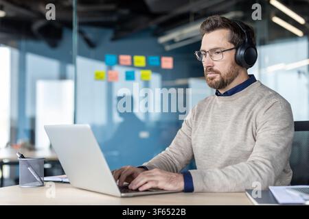 Businessman working on laptop and wearing headphones, focused on tasks at an office desk, illustrating remote work, concentration, or a virtual meeting Stock Photo