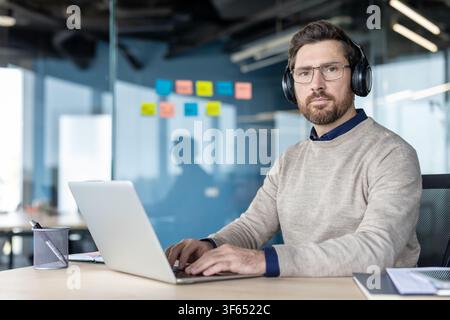Professional man wearing glasses and headphones, focused on working from a modern office, utilizing a laptop while engaging in virtual collaboration or deep concentration Stock Photo