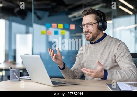 Happy smiling man wearing headphones and glasses, communicating during a virtual meeting or training session, working remotely from a modern office workspace Stock Photo