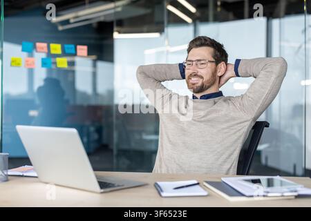 Young bearded man in glasses smiling and leaning back in his chair, enjoying a moment of success and accomplishment after completing work on his laptop in a modern office Stock Photo
