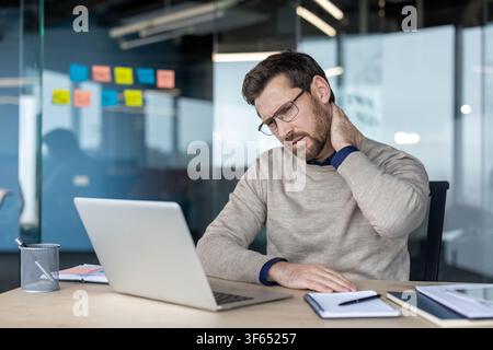Man feeling discomfort and rubbing his painful neck while sitting at a desk and using a laptop in a modern office environment, indicating stress, fatigue, and poor ergonomics at work Stock Photo