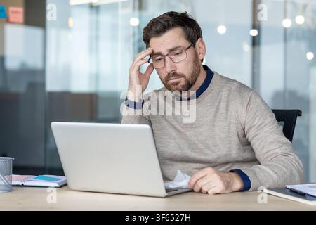 Mature businessman working at office desk with laptop, feeling tired and stressed, suffering from headache or burnout, holding a tissue Stock Photo